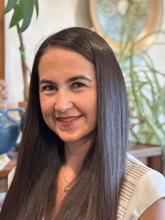 Smiling woman with long, straight dark hair wearing a sleeveless light-colored top, with plants and a decorative plate blurred in the background.