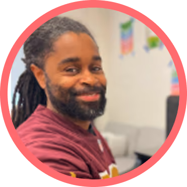 Close-up of a man with long dreadlocks and a beard smiling, wearing a maroon shirt inside a room with blurred background.
