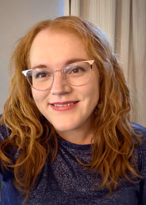 Smiling woman with curly red hair wearing clear-framed glasses and a dark blue glittery top, standing indoors in front of pale curtains.