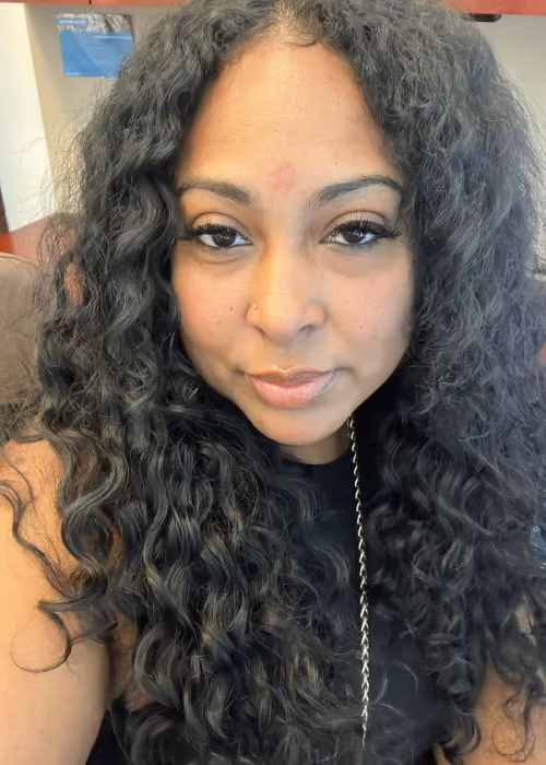 Close-up of a woman with long, curly black hair, natural makeup, and a nose piercing, wearing a black top and a necklace chain.