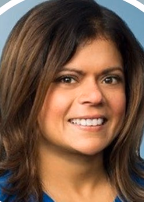 Smiling woman with medium-length brown hair wearing a blue top against a grayish-blue background.
