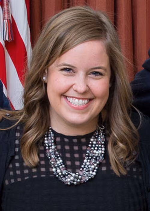 Smiling young woman with shoulder-length light brown hair wearing pearl earrings, a black top with a grid pattern, and a multi-strand beaded necklace, with a partially visible American flag and red curtain in the background.