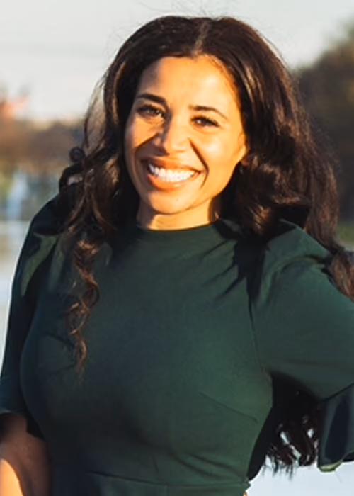 Smiling woman with long dark curly hair wearing a dark green top, standing outdoors with blurred trees and sky in the background.