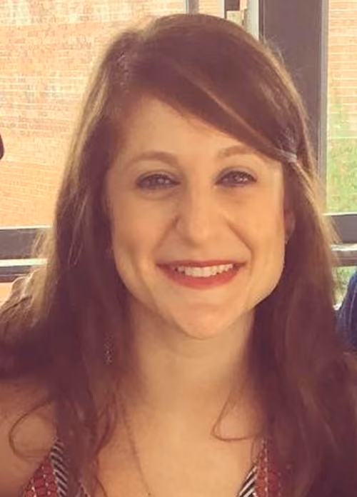 Smiling woman with long brown hair, wearing a patterned top, sitting indoors with a window and brick wall background.