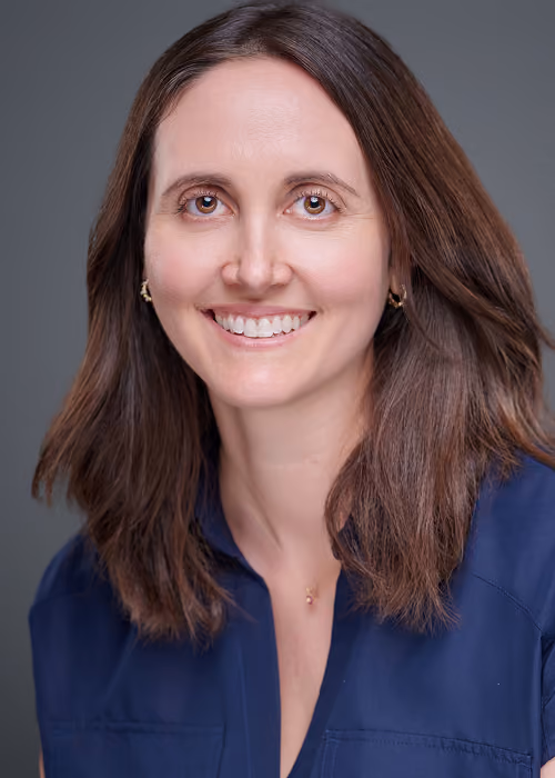 Smiling woman with shoulder-length brown hair wearing a dark blue top against a gray background.