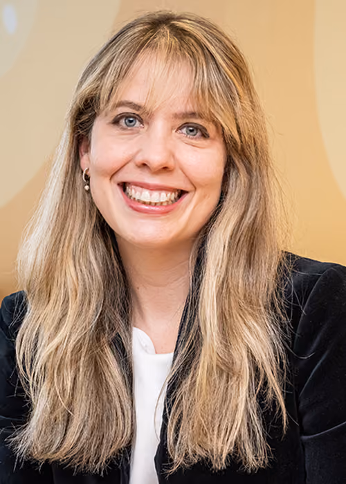 Smiling woman with long blonde hair wearing a black jacket and white top against a beige background.
