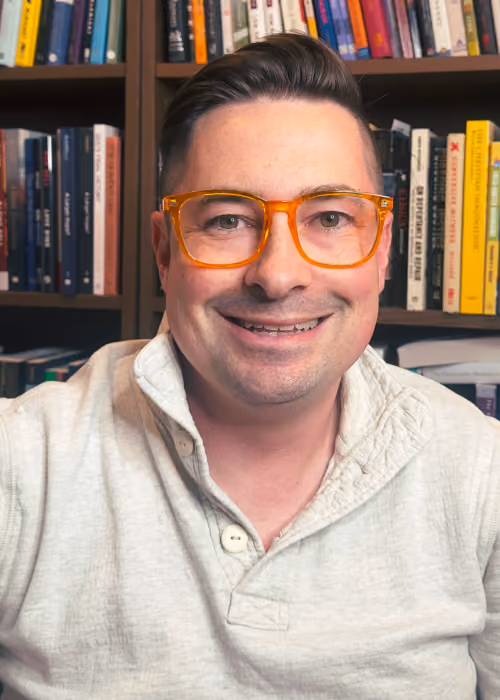 Smiling man wearing bright orange glasses and a light-colored sweater, sitting indoors with bookshelves filled with books in the background.