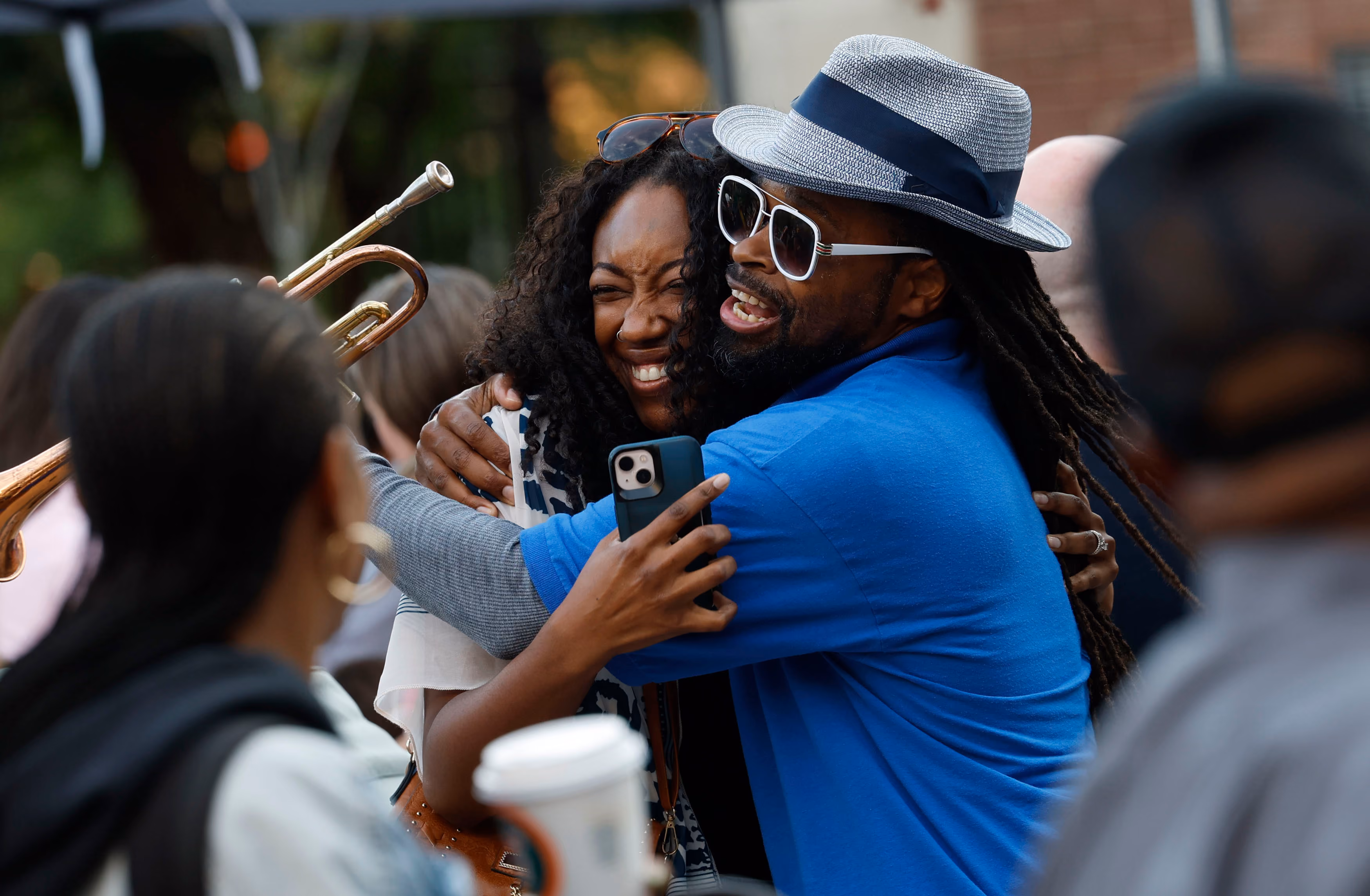 A man wearing a blue shirt, sunglasses, and a gray hat hugs a smiling woman with curly hair while holding a smartphone; two other people and a trumpet player are partially visible in the foreground and background.