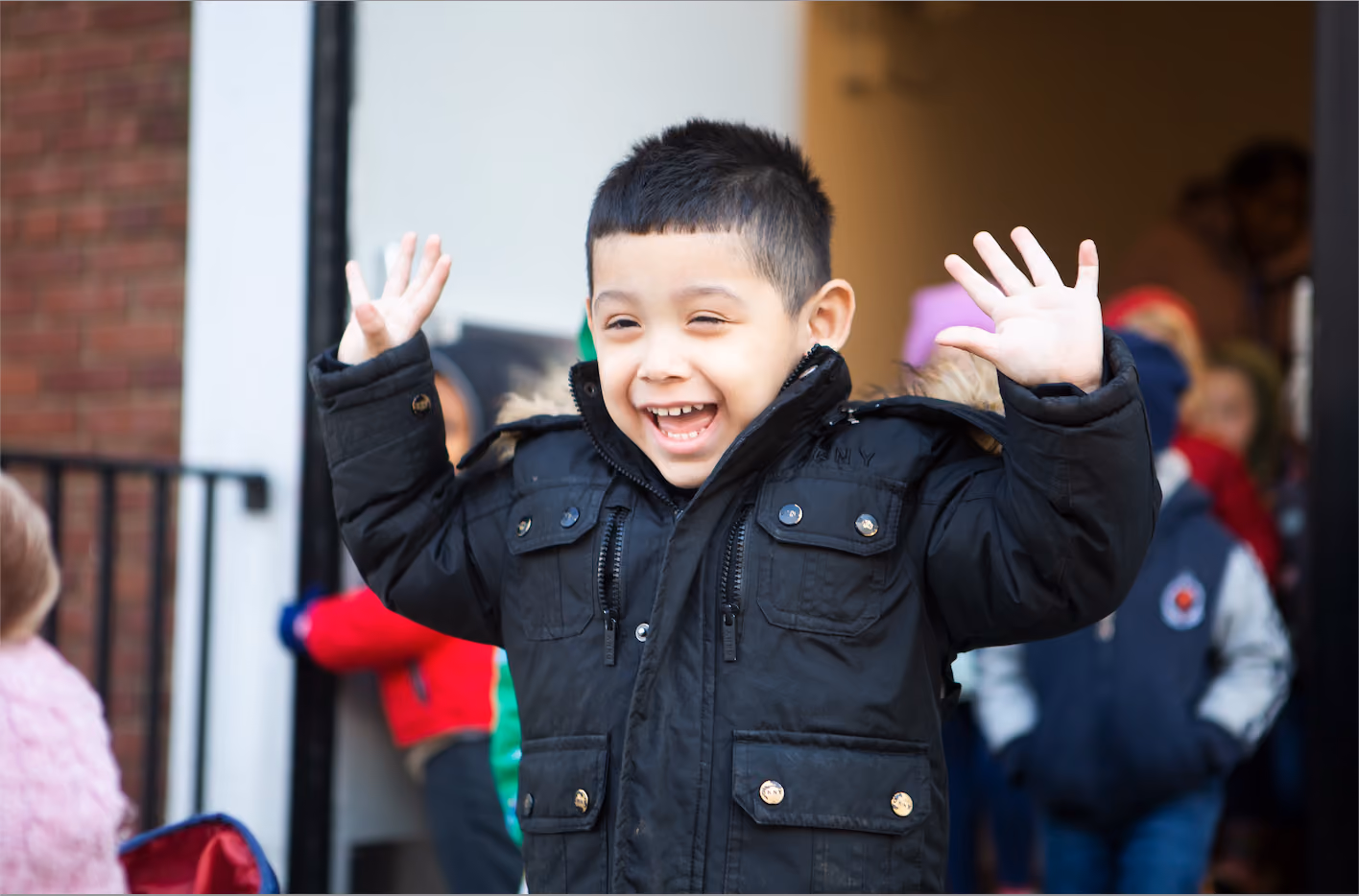 Smiling young boy wearing a black winter jacket with raised hands, standing outdoors near a building entrance with other children blurred in the background.