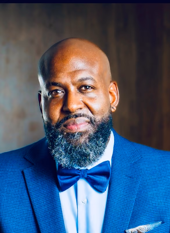 Portrait of a bald man with a full salt-and-pepper beard wearing a blue checkered suit, white shirt, and a blue bow tie, smiling slightly against a blurred brown background.