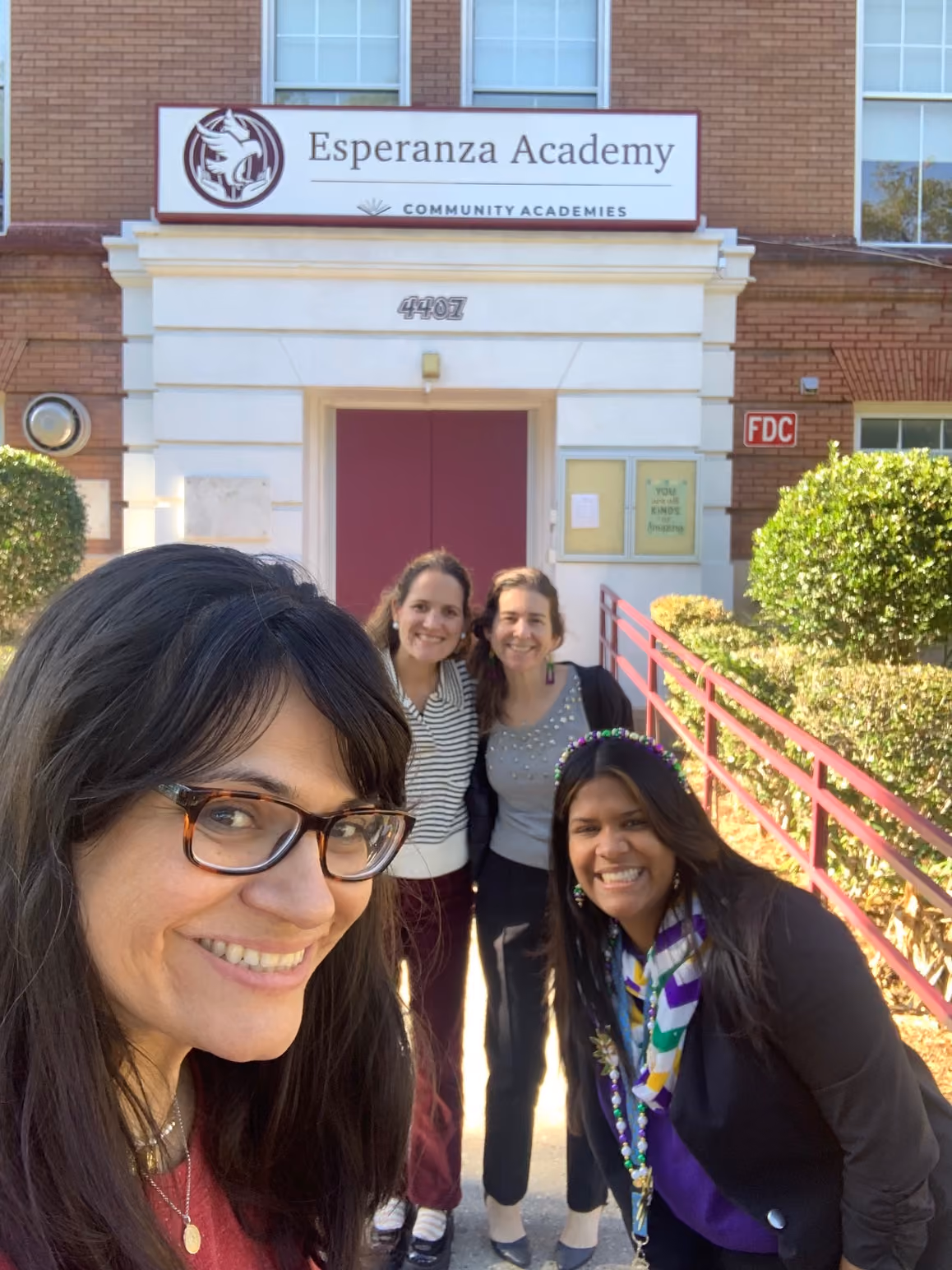 Four smiling women posing for a selfie outside the entrance of Esperanza Academy, a brick building with a sign above the door reading 'Esperanza Academy, Community Academies.'