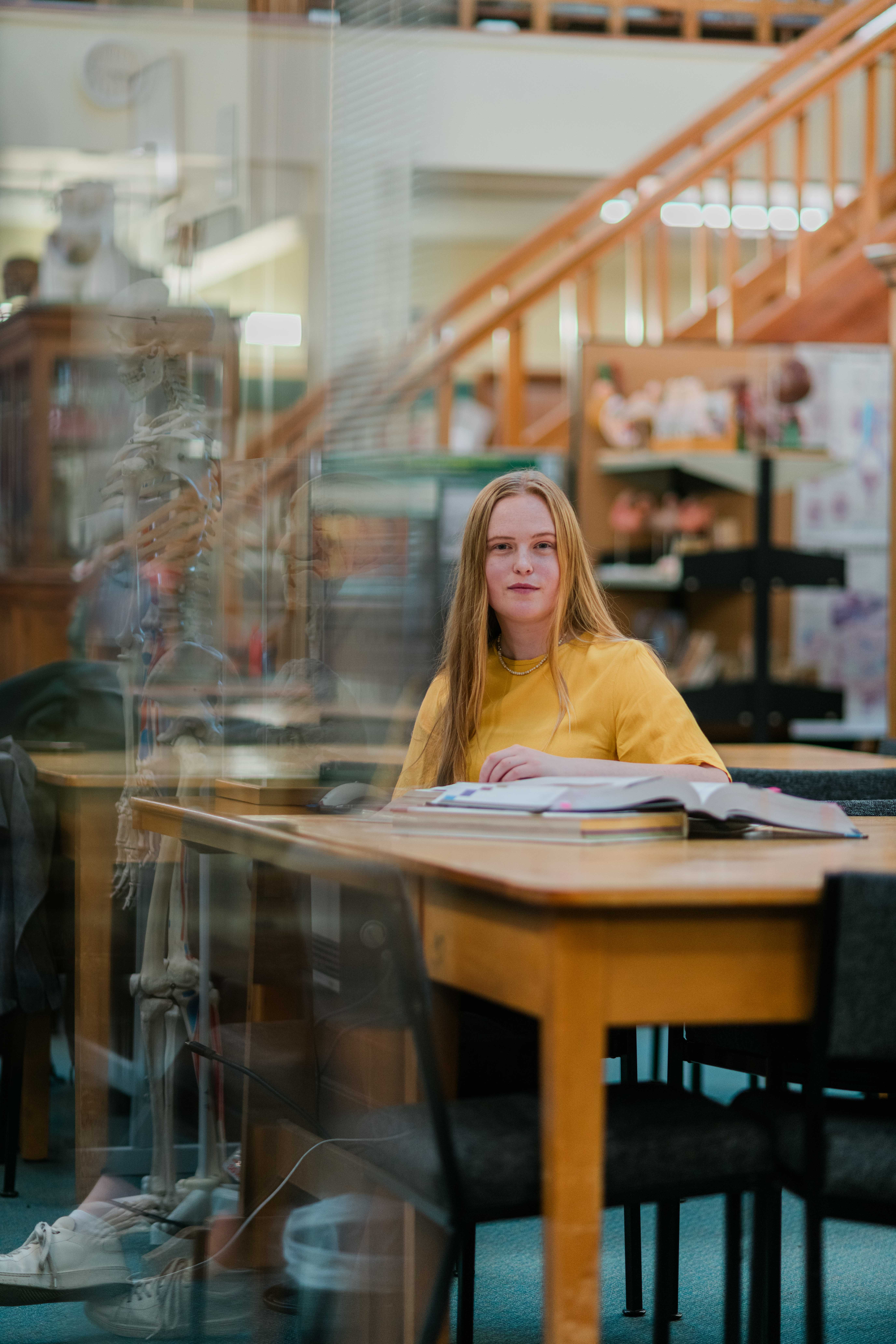 Young woman in a yellow shirt sitting at a library table with open books, with a skeleton model reflected in glass nearby.