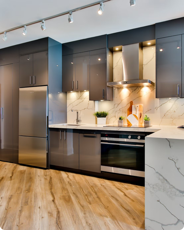 Modern kitchen with gray cabinets, stainless steel fridge, oven, marble countertops, and wooden flooring.