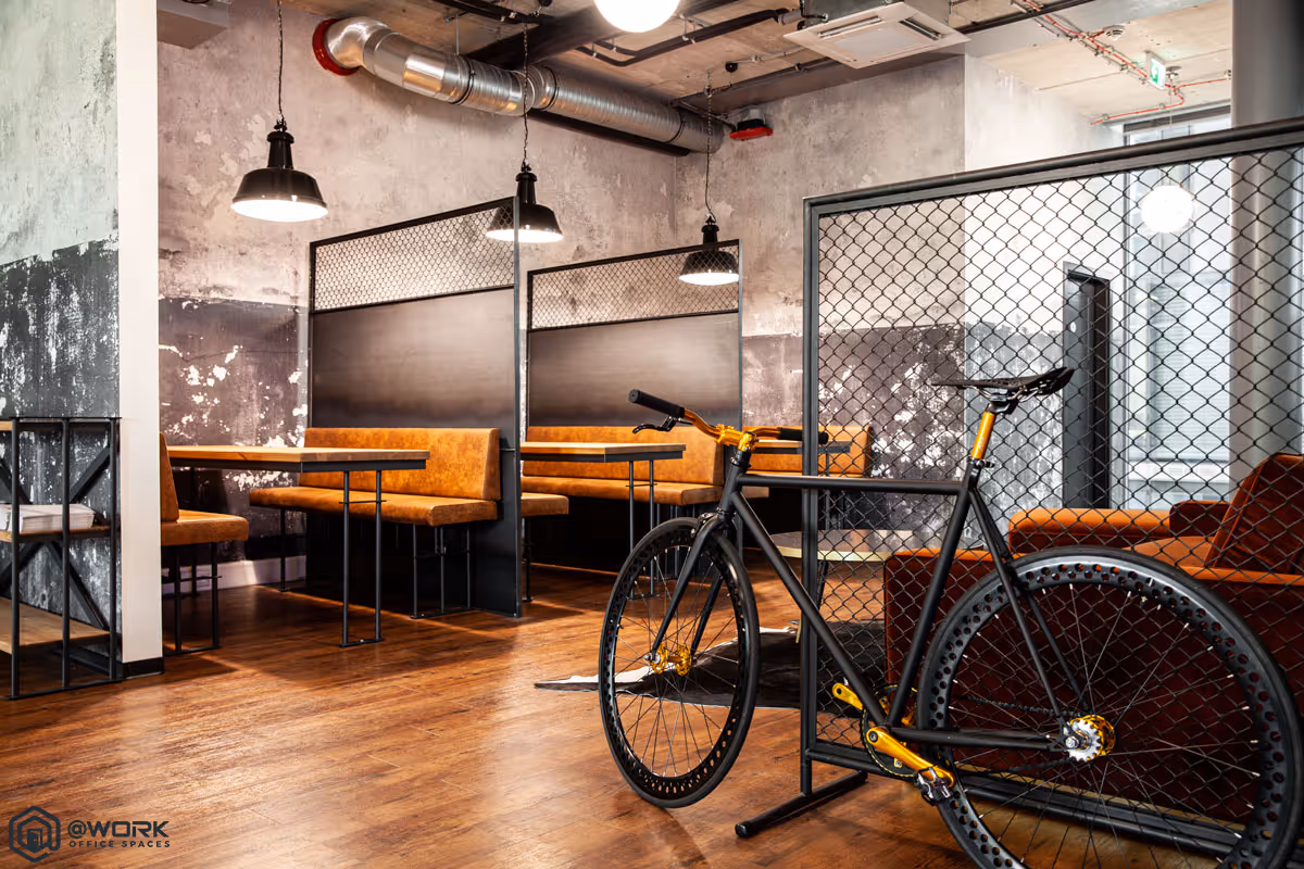 Modern office lounge with wooden floors, brown leather booth seating, industrial hanging lights, and a black bicycle with golden accents behind a wire mesh partition.