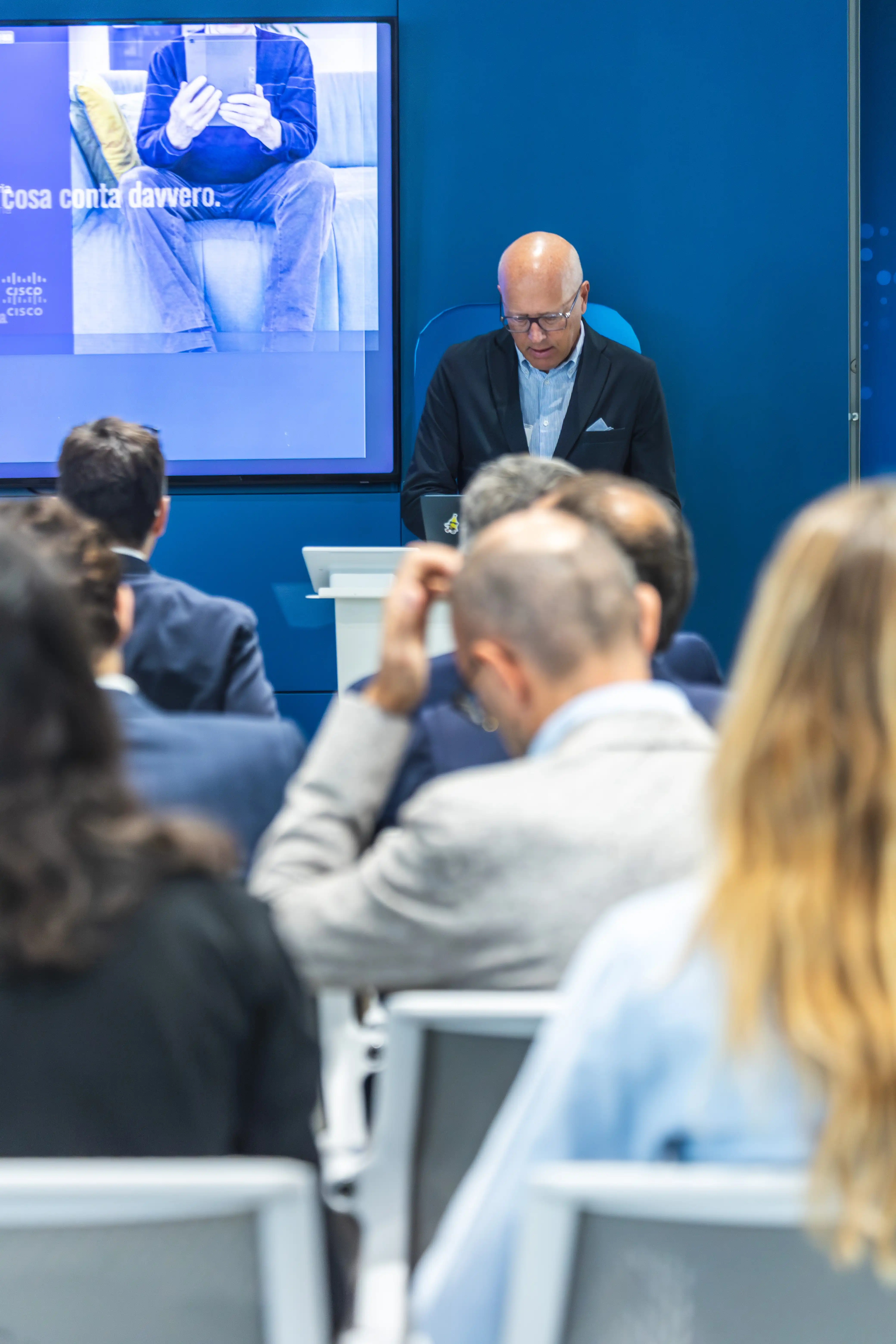 Uomo in giacca scura che parla davanti a un pubblico seduto durante una presentazione in una sala con pareti blu.