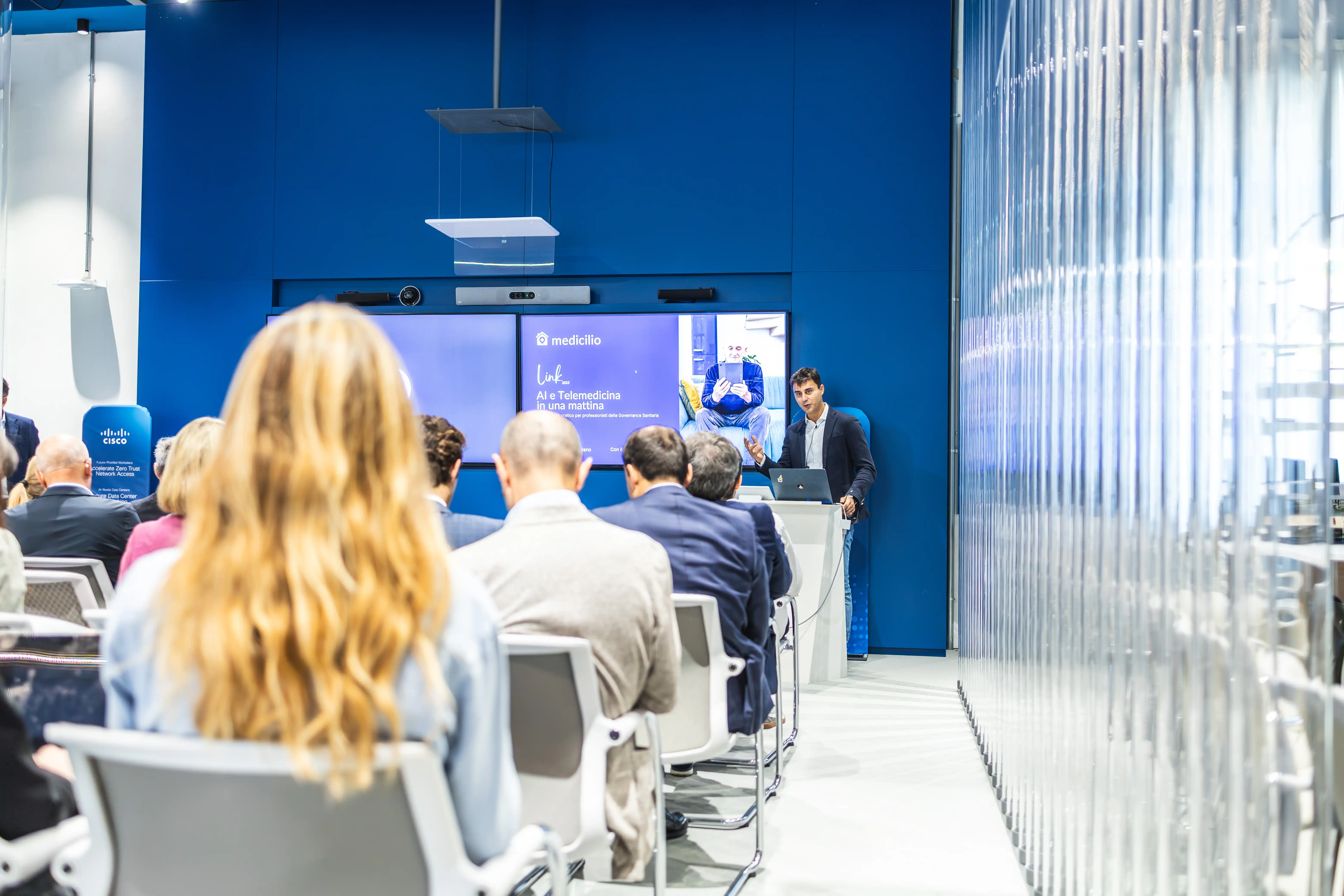 Un uomo in abito scuro presenta una conferenza su telemedicina davanti a un pubblico seduto in una sala con pareti blu e schermi digitali.
