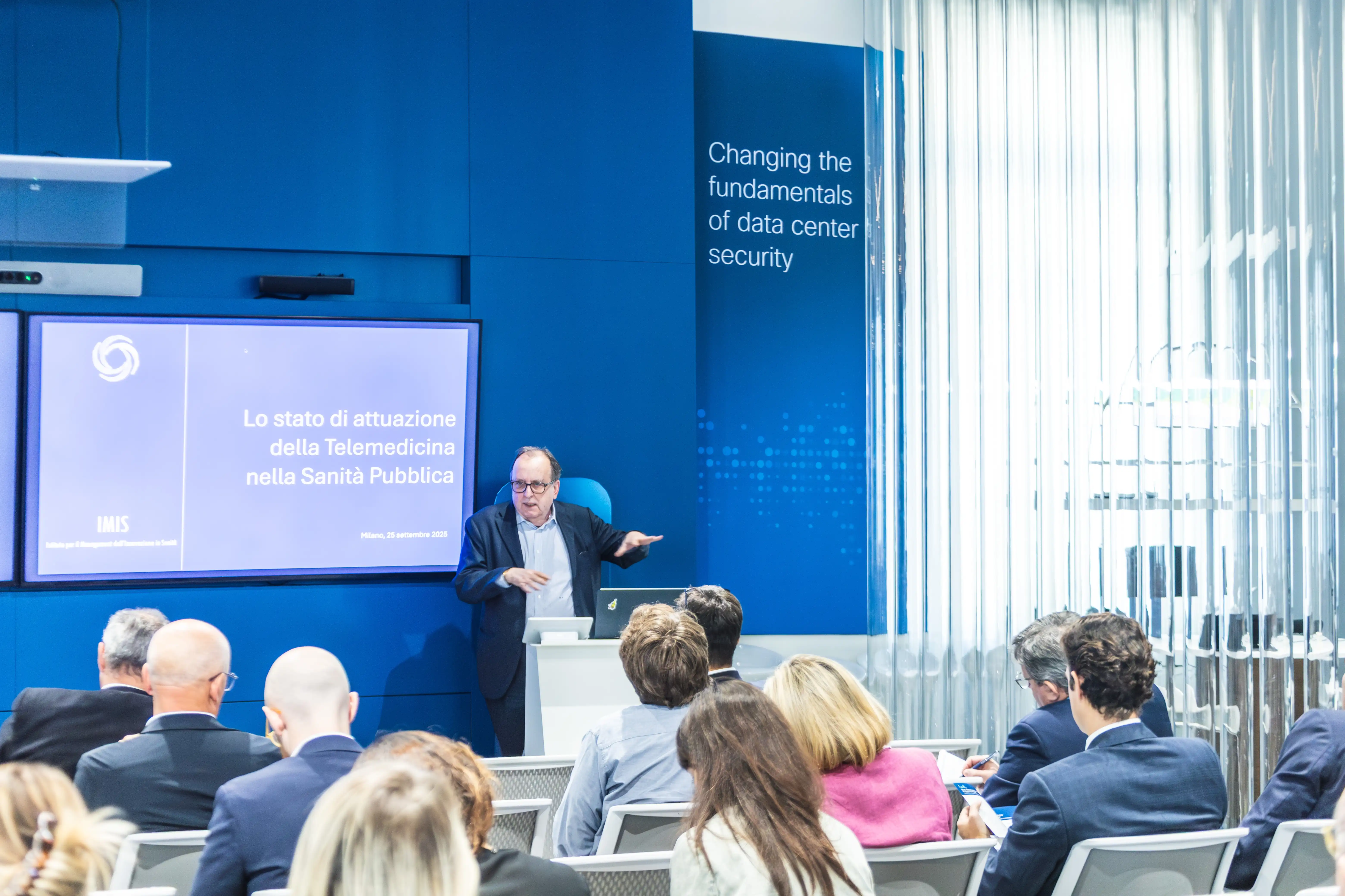 Un uomo in giacca che tiene una presentazione sulla telemedicina nella sanità pubblica davanti a un pubblico seduto in una sala conferenze con pareti blu.