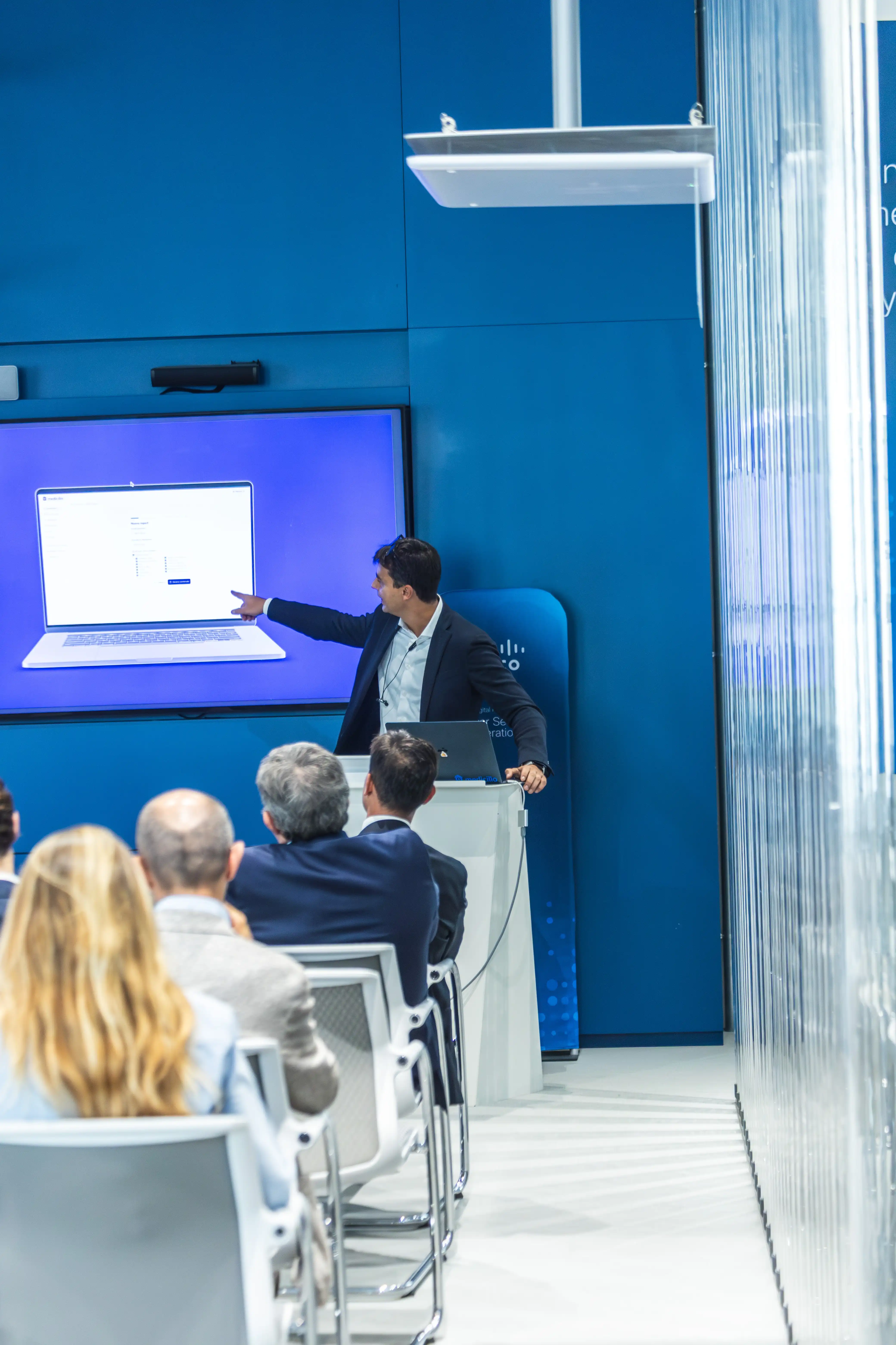 Mattia Perroni che presenta a un pubblico seduto, indicando un grande schermo con immagine di laptop.