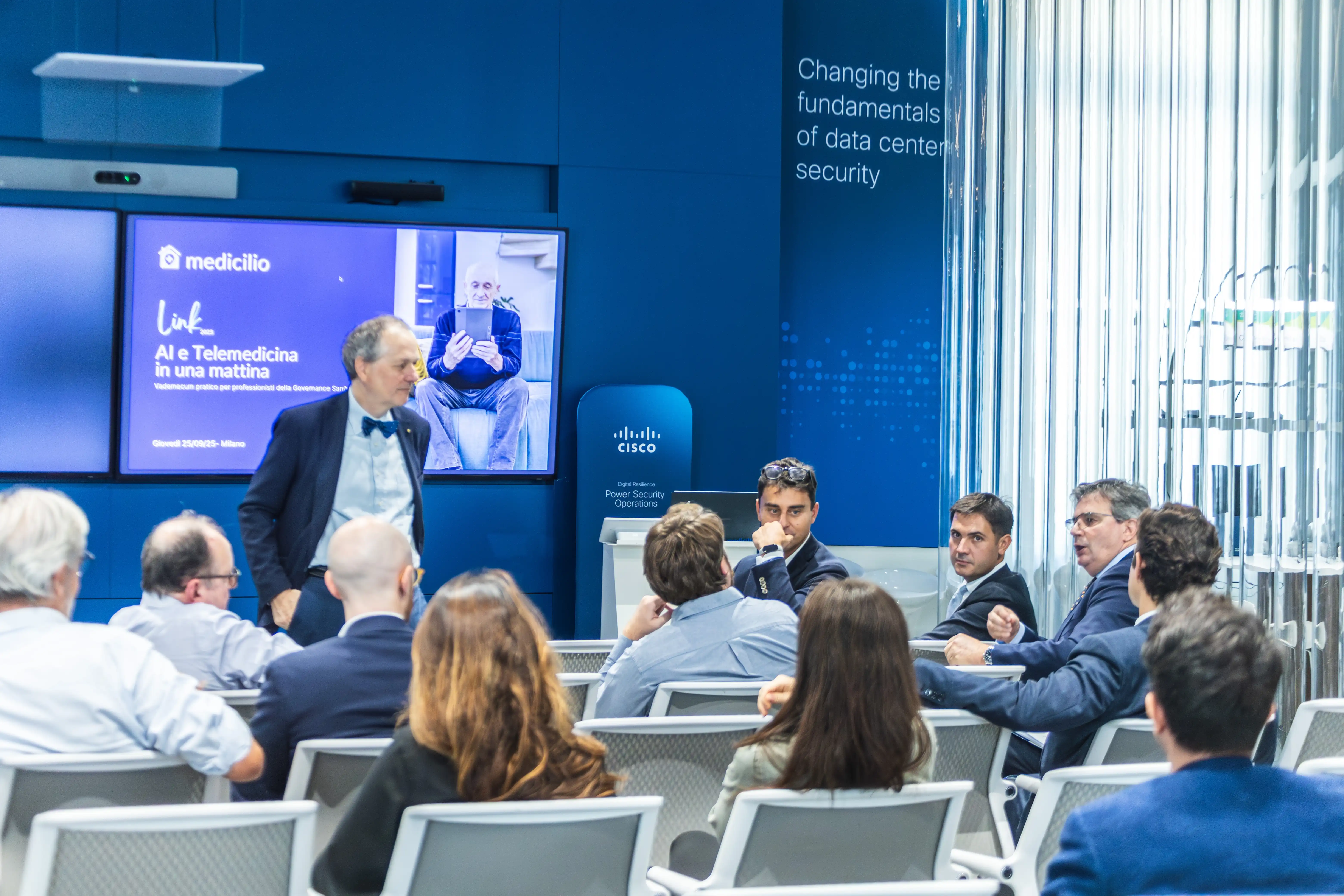 Uomo in giacca blu che parla davanti a un pubblico seduto in una sala conferenze con schermo che mostra presentazione su AI e telemedicina a Milano.