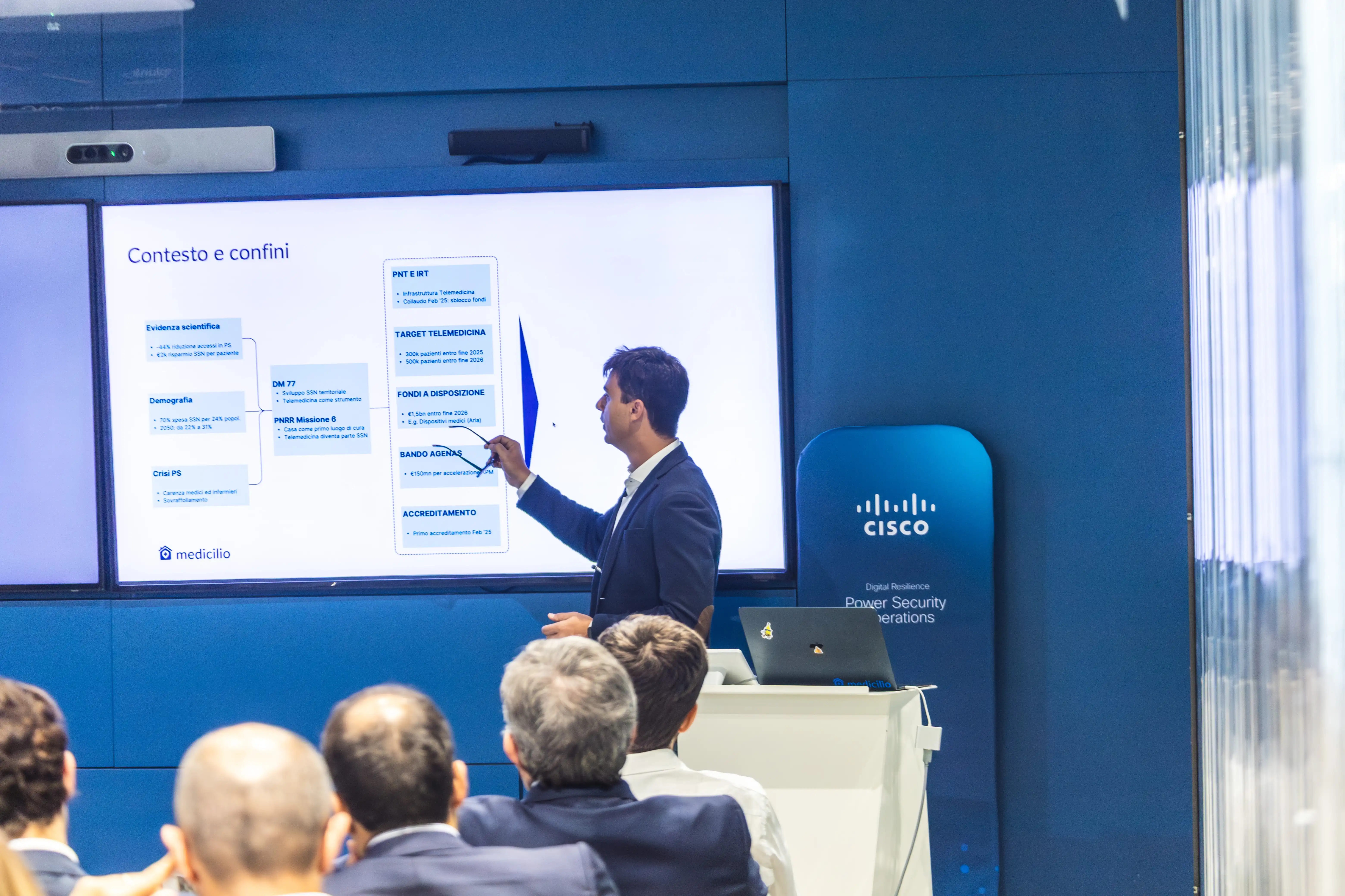 Uomo in abito che fa una presentazione davanti a uno schermo con diagramma chiamato Contesto e confini in una sala conferenze con pubblico.