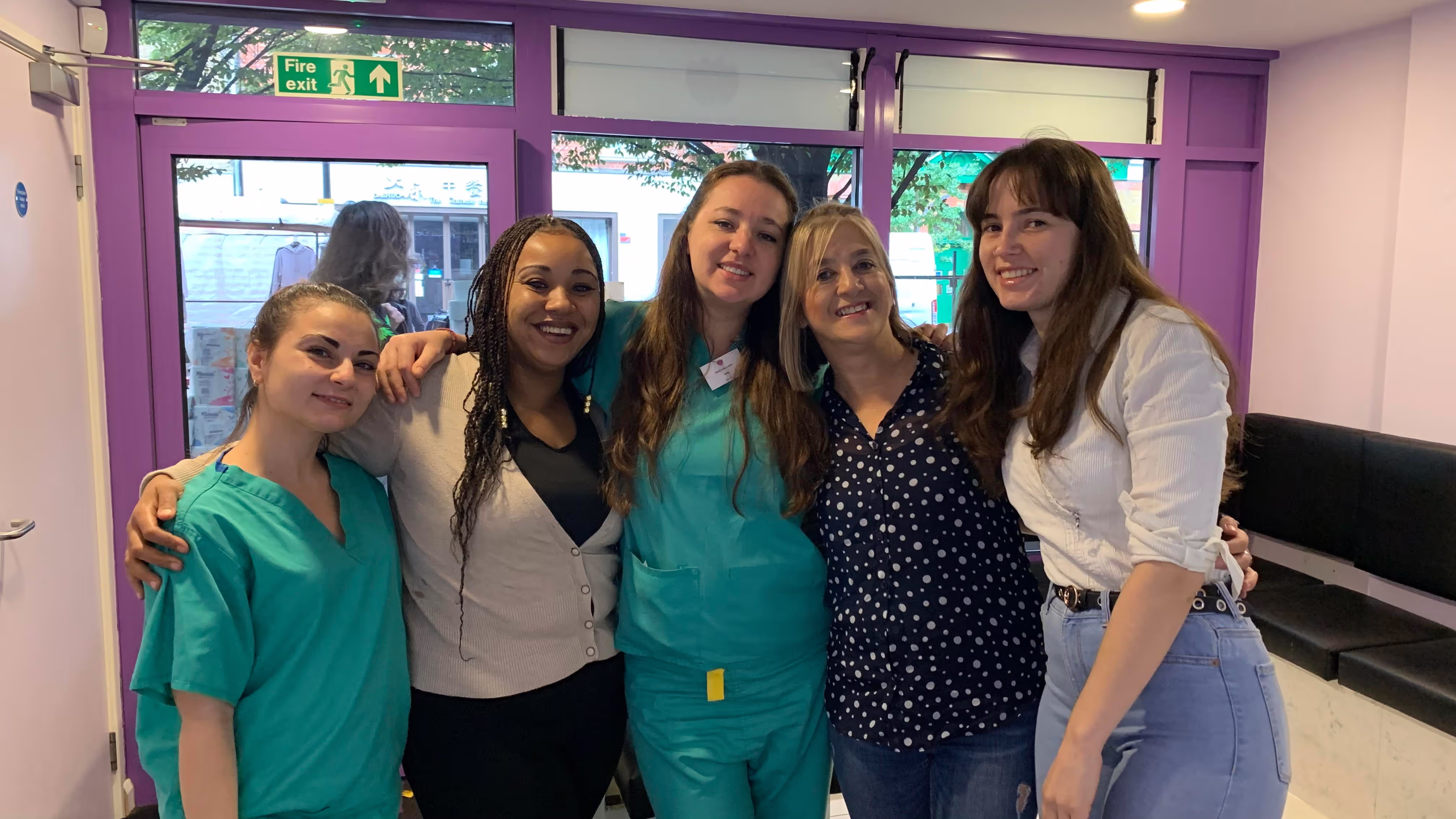 Group of five smiling women standing closely together in a room with purple doors and windows in the background.