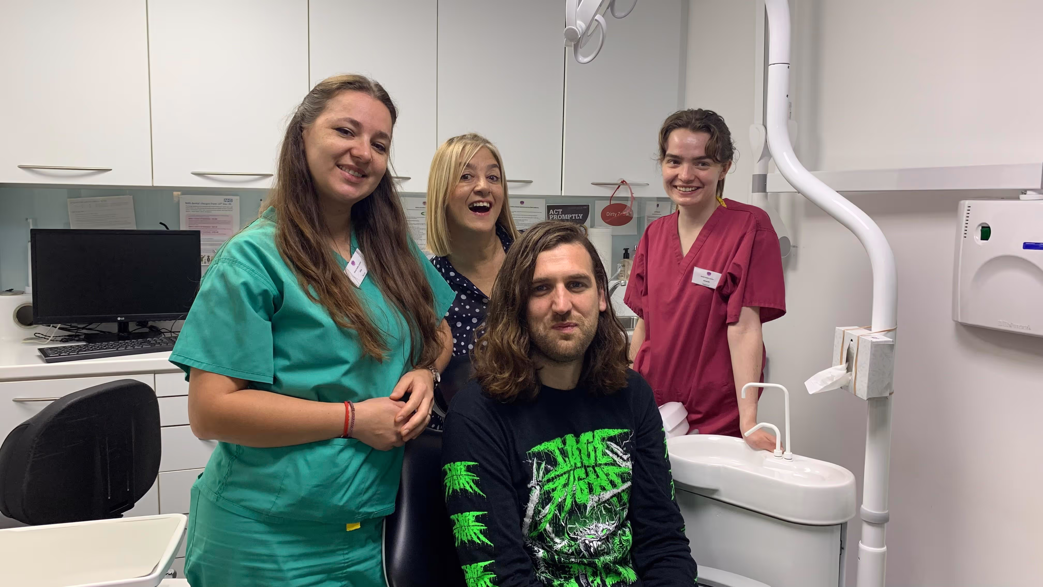 Three smiling dental staff members in scrubs posing with a male patient sitting in a dental chair in a clinic room.