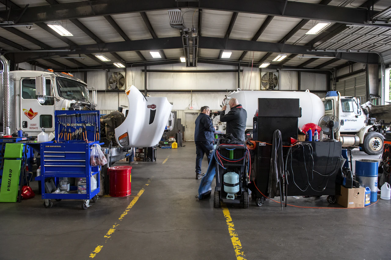 Inside a well-lit truck maintenance shop, two workers converse between service bays where multiple trucks with raised hoods are undergoing repairs. The space is organized with tool carts, equipment, and supplies neatly arranged along yellow-striped work zo