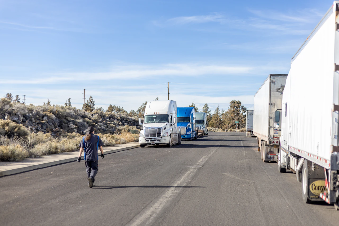 A person in a dark work uniform walks down a wide asphalt road toward a row of parked semi-trucks, including a white and a blue tractor-trailer, under a clear blue sky. The scene is set in a dry, rural area with scattered shrubs and rocky terrain.