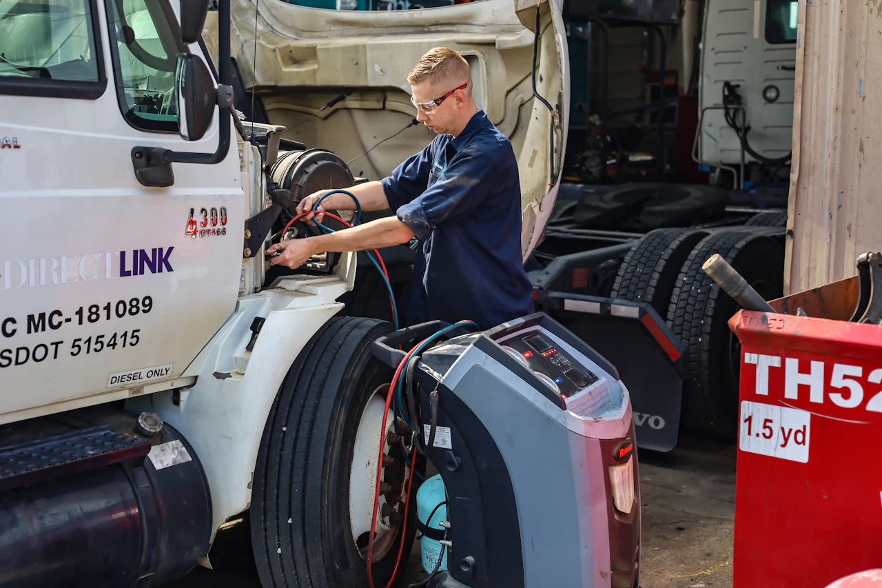 A mechanic wearing safety glasses works on a white semi-truck with its hood raised, using diagnostic equipment connected by red and blue hoses. The scene takes place in a busy garage with tools, tires, and a red waste bin labeled “TH52” nearby.