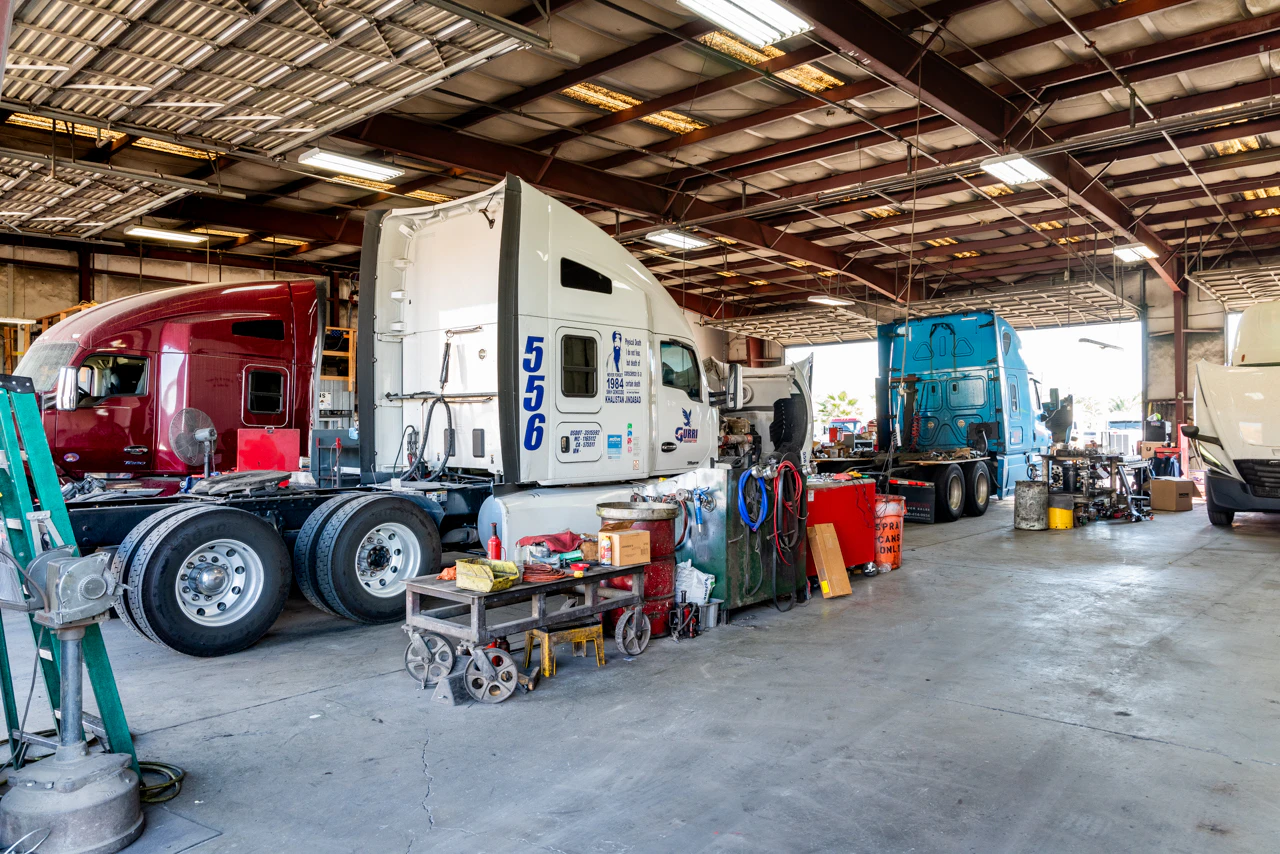 Several semi-trucks in red, white, blue, and white are lined up inside a spacious repair garage, with their hoods open and maintenance tools scattered on workbenches. The facility is well-lit with fluorescent lights and has a high, open-beam ceiling.