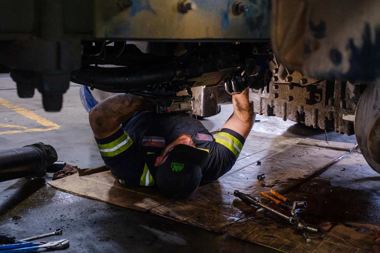 A mechanic lies beneath a heavy-duty truck, using tools to work on the drivetrain components. The scene shows the undercarriage, with oil stains and wrenches nearby, highlighting the hands-on nature of truck repair work.
