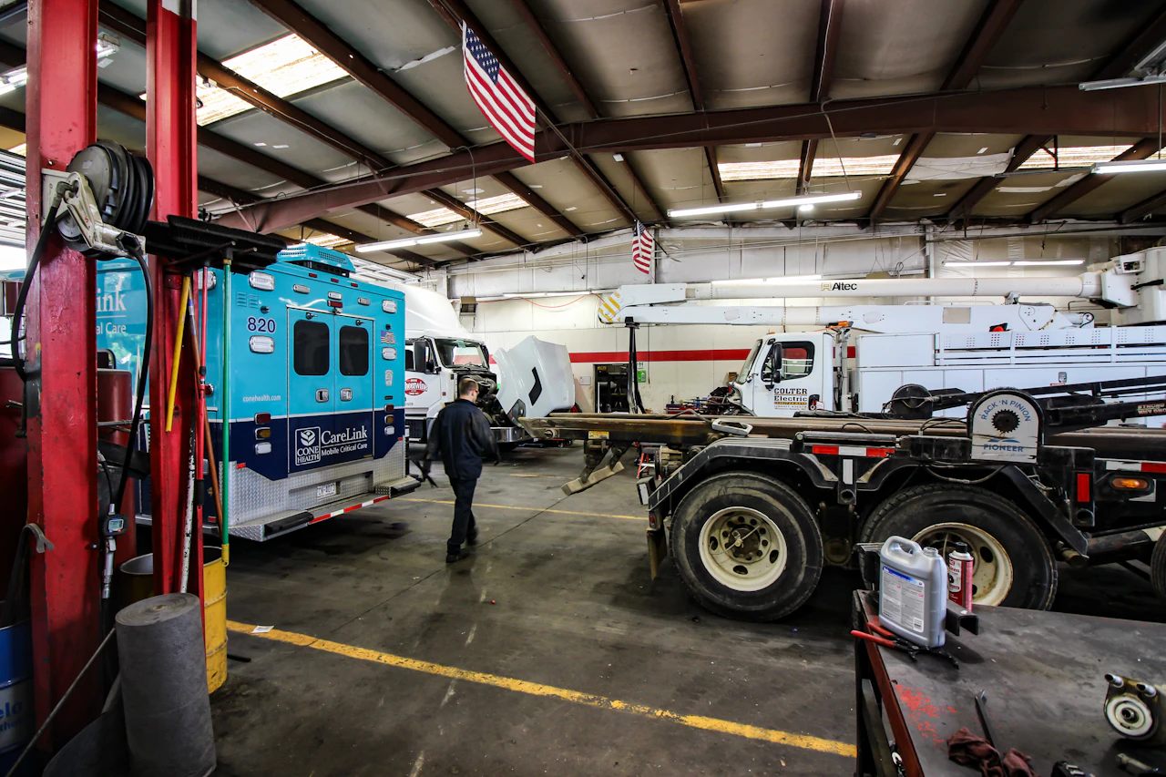 Inside a vehicle maintenance garage, a technician walks past a teal CareLink mobile critical care ambulance, with large trucks, a utility vehicle, and a semi-truck . The space is organized with tools, equipment, and American flags hanging from the ceiling.