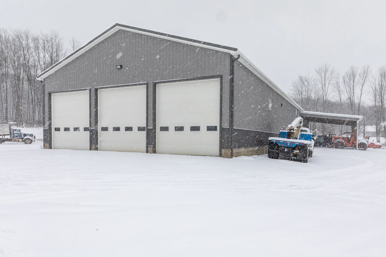 Alt Text Generator said: A gray industrial garage with three large white bay doors stands in a snowy landscape as snow falls steadily; nearby, a blue tow truck and orange tractor are parked by the side and under a shelter.