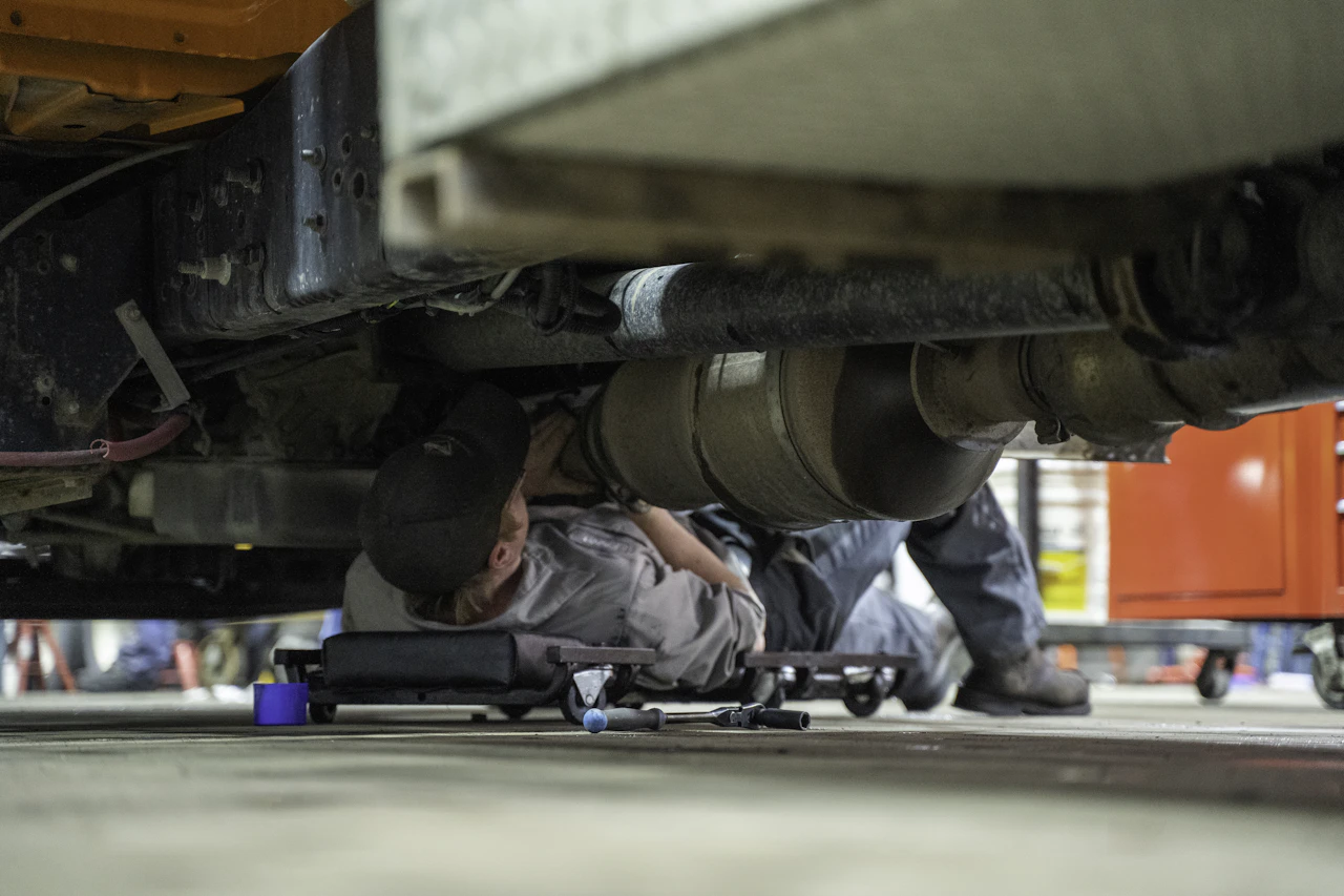 A mechanic lies on a creeper under a large vehicle, working near the exhaust system in a well-lit garage. Tools and a red tool chest are visible nearby, indicating an active repair environment.
