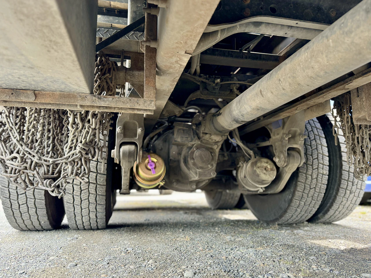 Close-up view under a heavy-duty truck shows the drivetrain, axles, and dual rear wheels, with tire chains hanging and a gold brake chamber featuring a purple release valve.