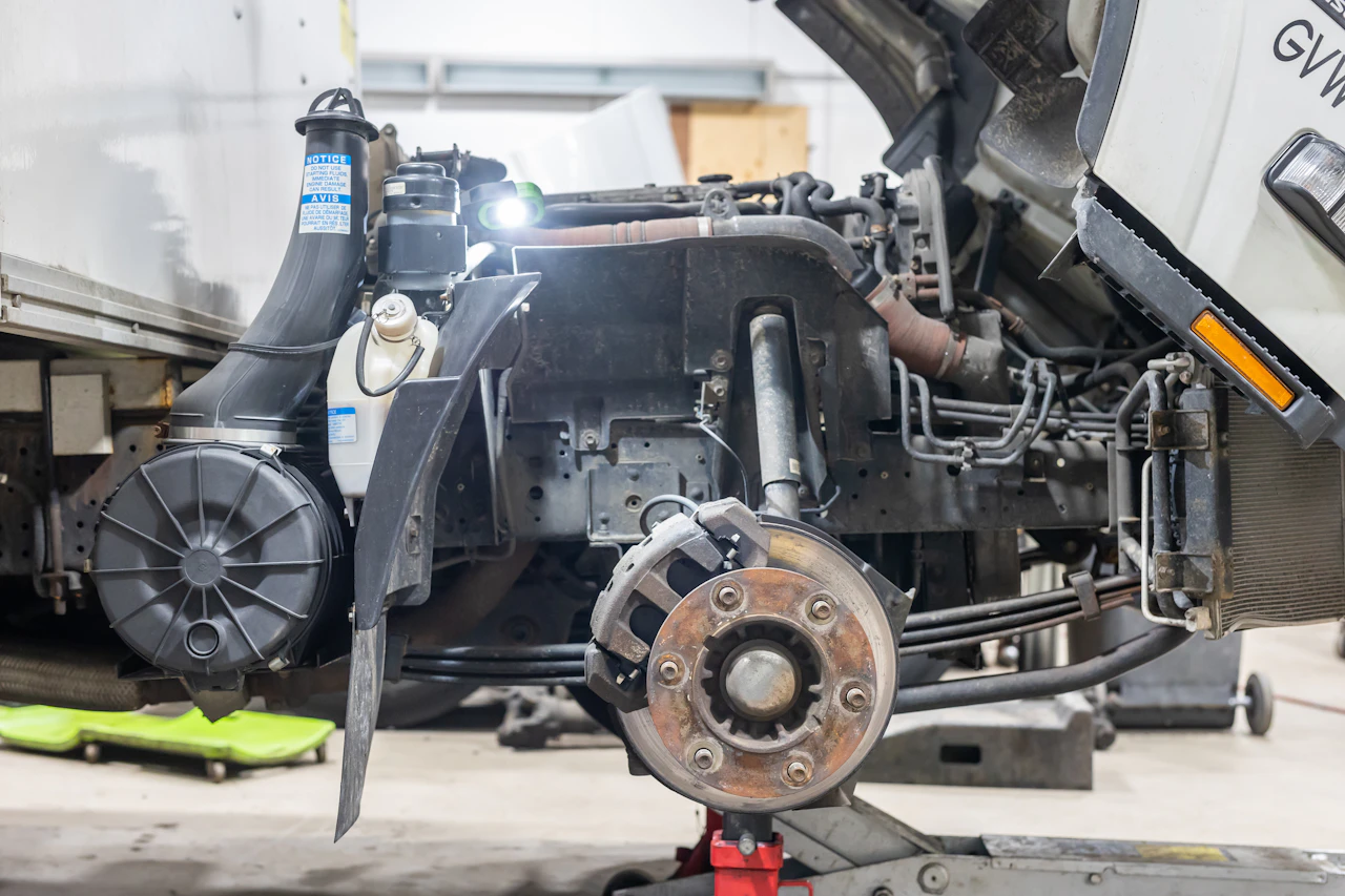 A semi-truck with its front wheel removed is propped on a jack, exposing the brake rotor, suspension, and engine components. A bright work light illuminates the mechanical parts inside the open hood.