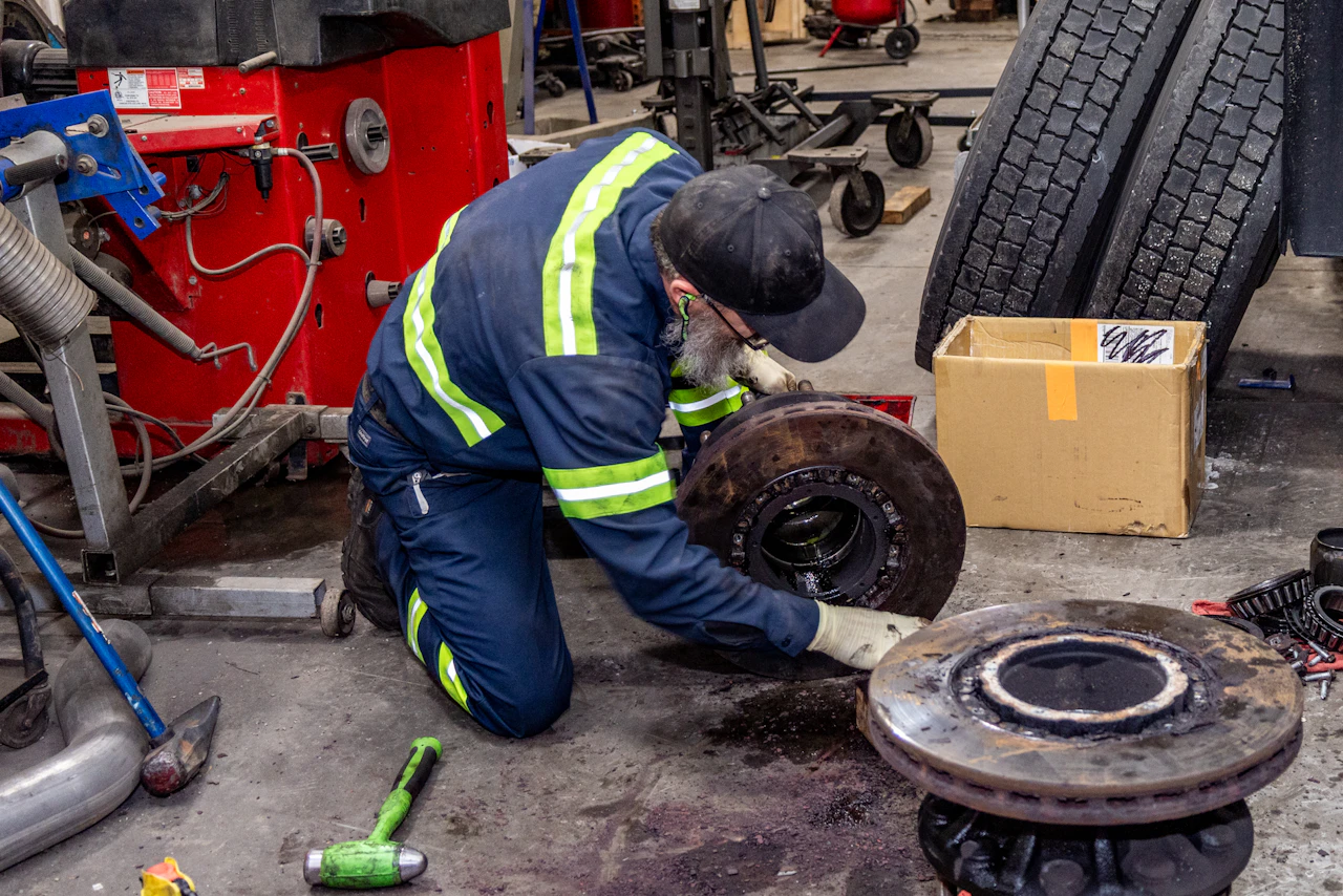 A mechanic in a navy uniform with reflective stripes kneels on a garage floor while repairing a large brake assembly. Heavy truck tires, tools, and a red machine surround him in a busy auto shop.