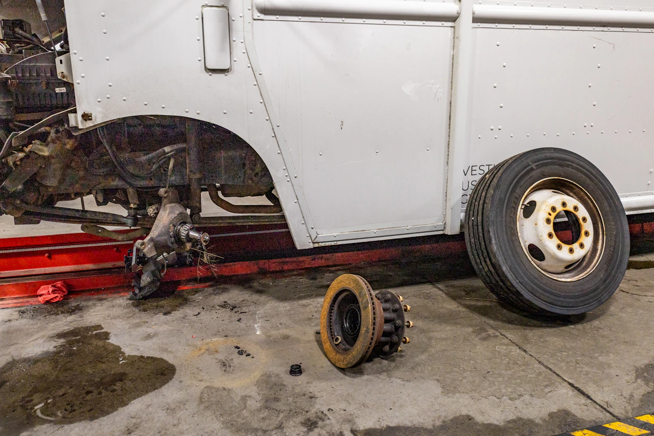 A white service truck sits lifted in a repair shop with its rear wheel removed, exposing the axle and brake components. A large brake drum rests on the stained concrete floor beneath the vehicle.