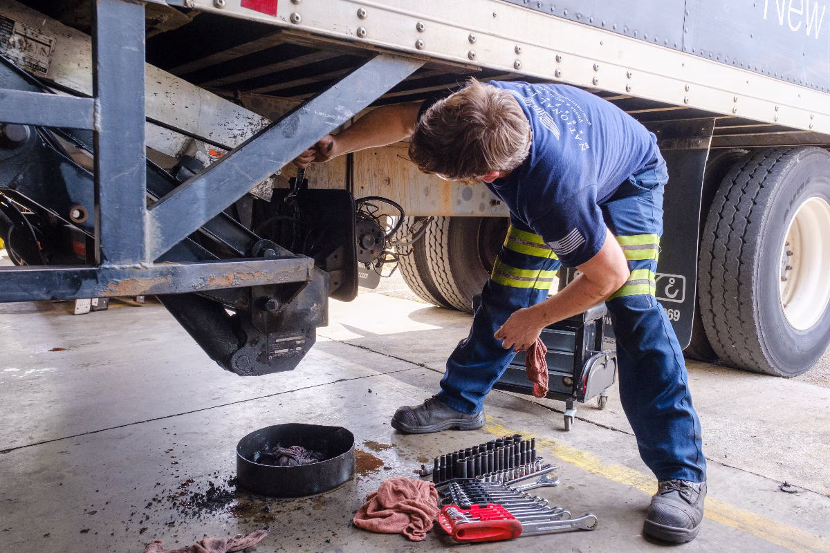 Mechanic repairing trailer undercarriage using tools with oil pan and rags on ground nearby workshop