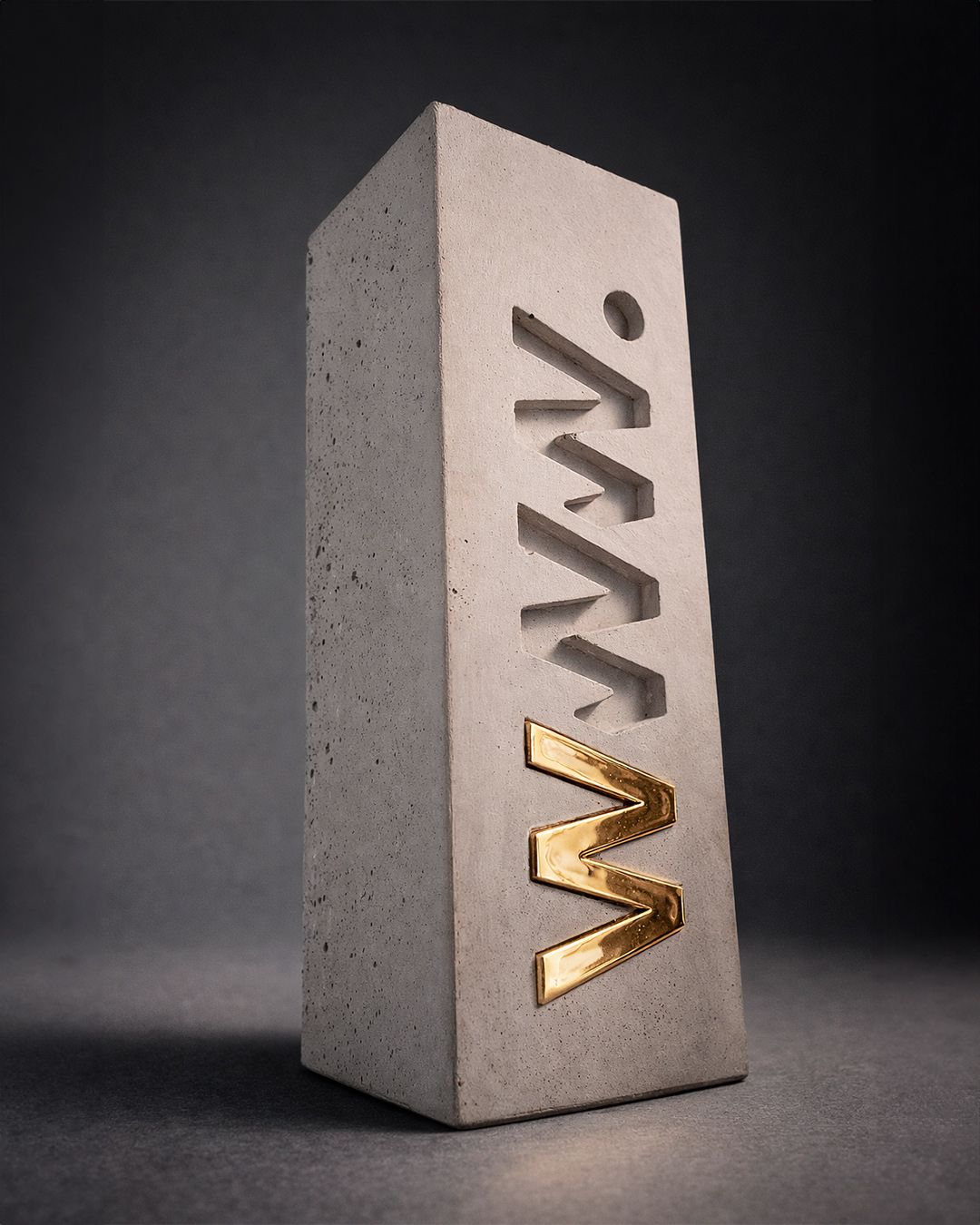 Tall rectangular concrete block with the acronym 'MWM' carved vertically and a gold metallic 'M' attached at the bottom.