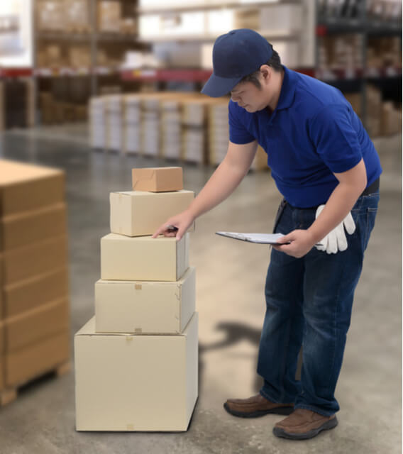 Warehouse worker in a blue uniform checking stacked delivery boxes with a clipboard, representing inventory and product management services.