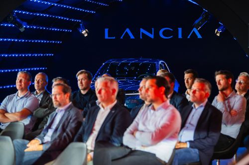 Audience of men seated in a dark room with blue LED lighting and a Lancia car displayed behind them.