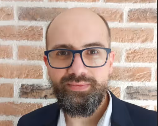 Bald man with a beard and glasses wearing a dark blazer and white shirt, standing against a brick wall.