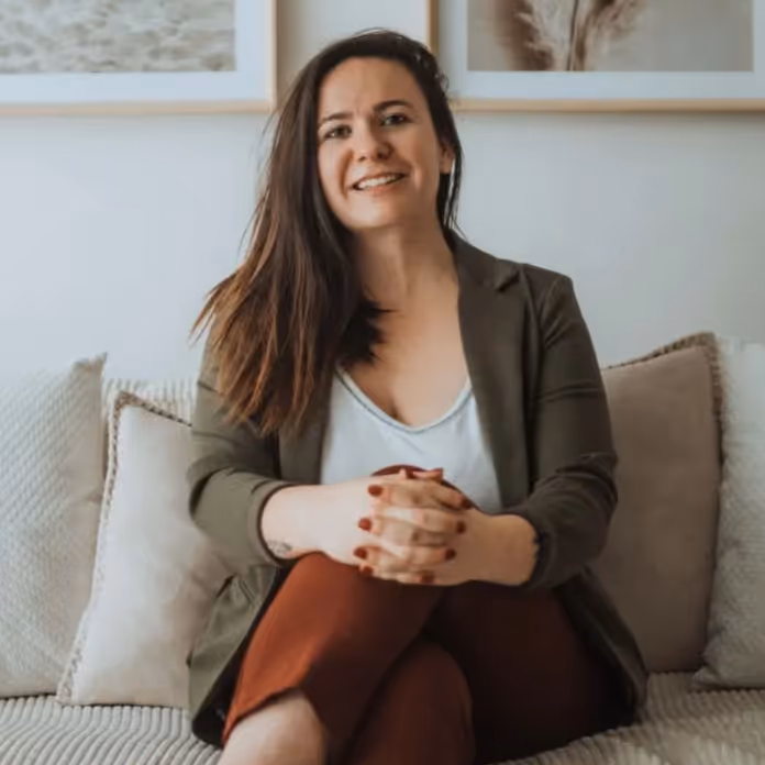 Smiling woman with long dark hair sitting cross-legged on a sofa with neutral pillows and wall art behind her.