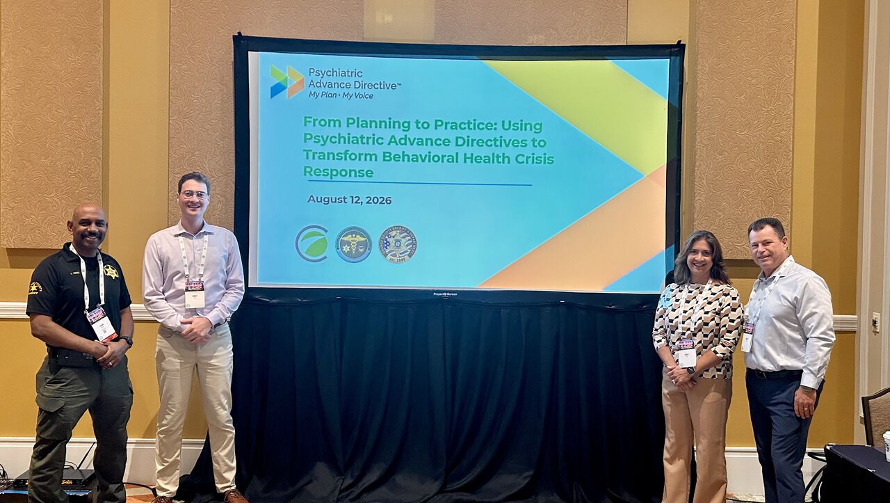 An image of members of Orange County Sheriff's department and Concepts Forward Consulting standing in front of a screen showing the title slide of their PowerPoint presentation. The title slide says "From Planning to Practice: Using Psychiatric Advance Directives to Transform Behavioral Health Crisis Response"