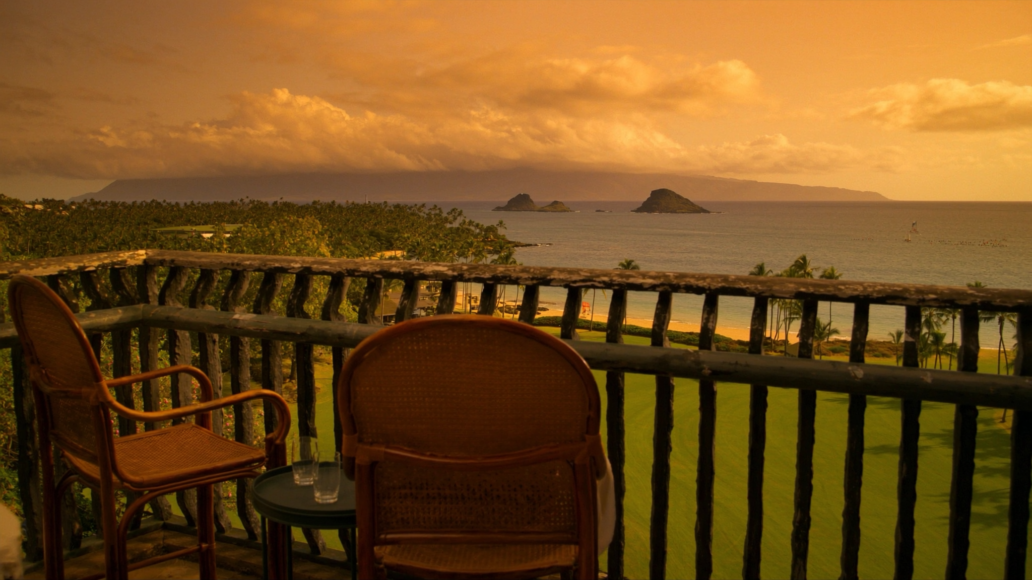Two empty chairs on a private oceanfront lanai at The Hale Pau Hana in Kihei, looking across the lawn and the sand of Kamaole Beach Park II toward Lanai and Kahoolawe at sunset