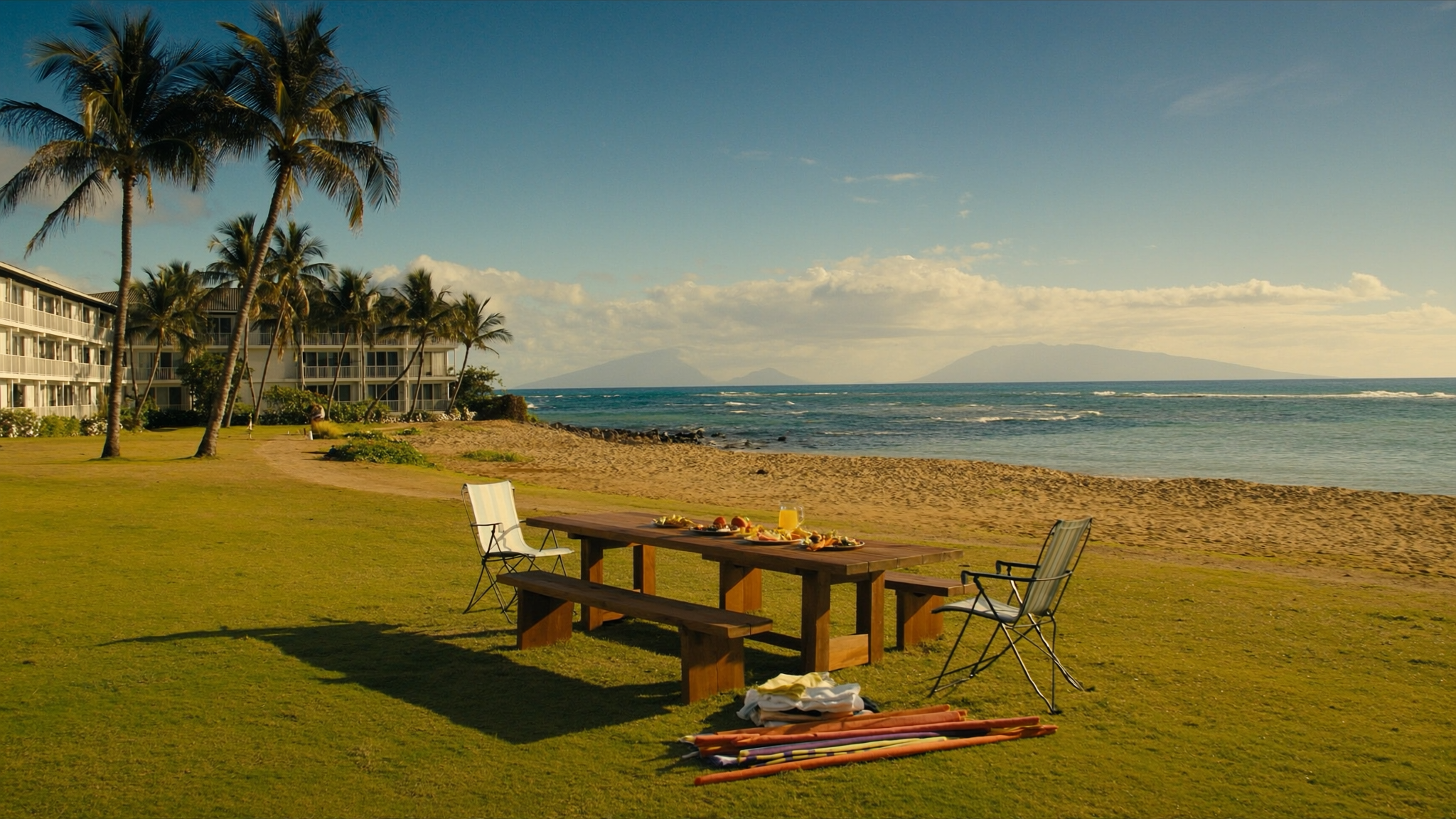 A long family table set for a shared meal on the oceanfront lawn where The Hale Pau Hana meets the sand of Kamaole Beach Park II in Kihei, Maui