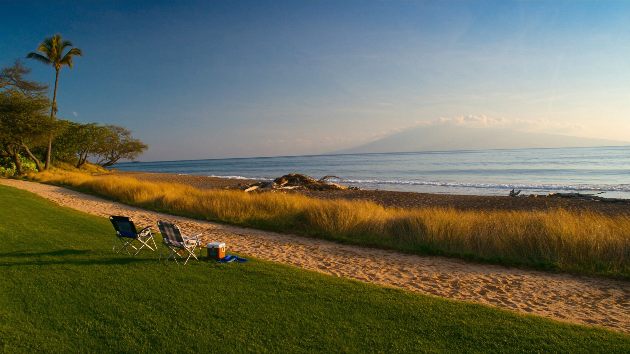 The oceanfront lawn at The Hale Pau Hana meeting the sand of Kamaole Beach Park II in Kihei, Maui, with beach chairs and a cooler staged on the grass