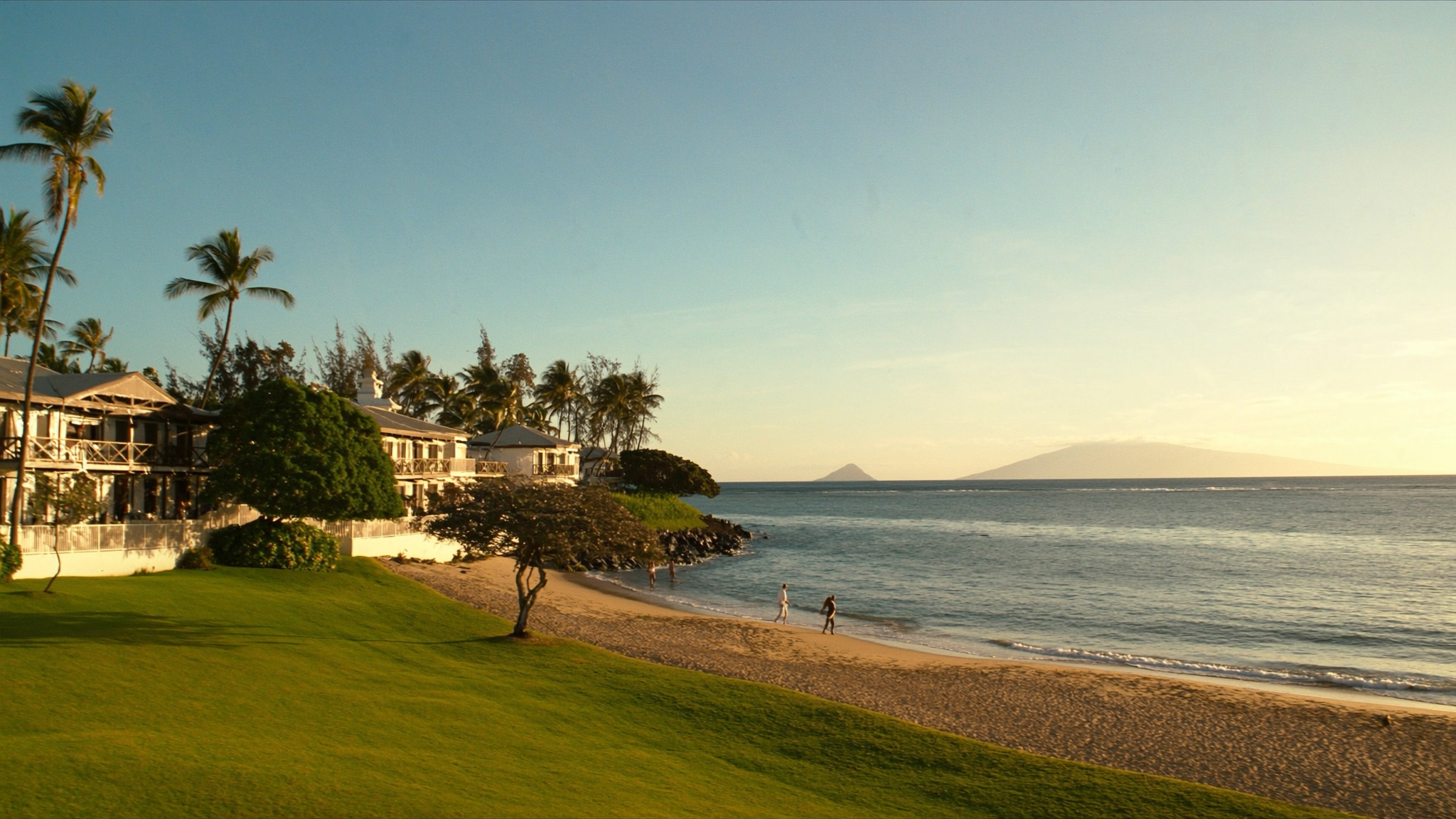 The oceanfront lawn at The Hale Pau Hana meeting the sand of Kamaole Beach Park II in Kihei, Maui, with guests in aloha attire walking the shoreline at golden hour