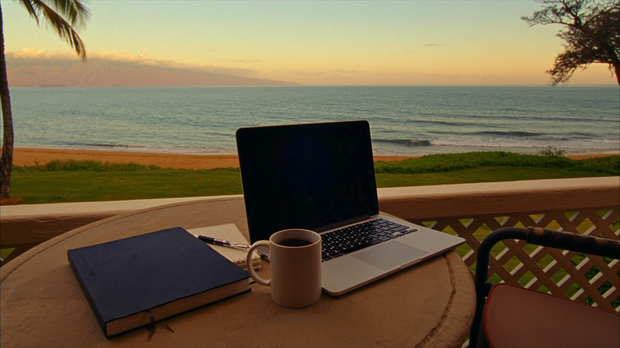 A laptop and a mug of coffee on a private oceanfront lanai in Kihei, Maui, looking out over the lawn and the sand of Kamaole Beach Park II in the early morning